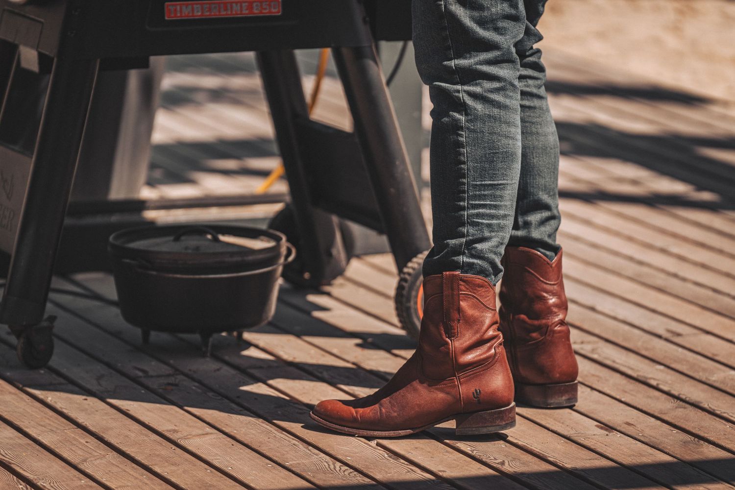 Person wearing brown leather cowboy boots and jeans standing on a wooden deck.