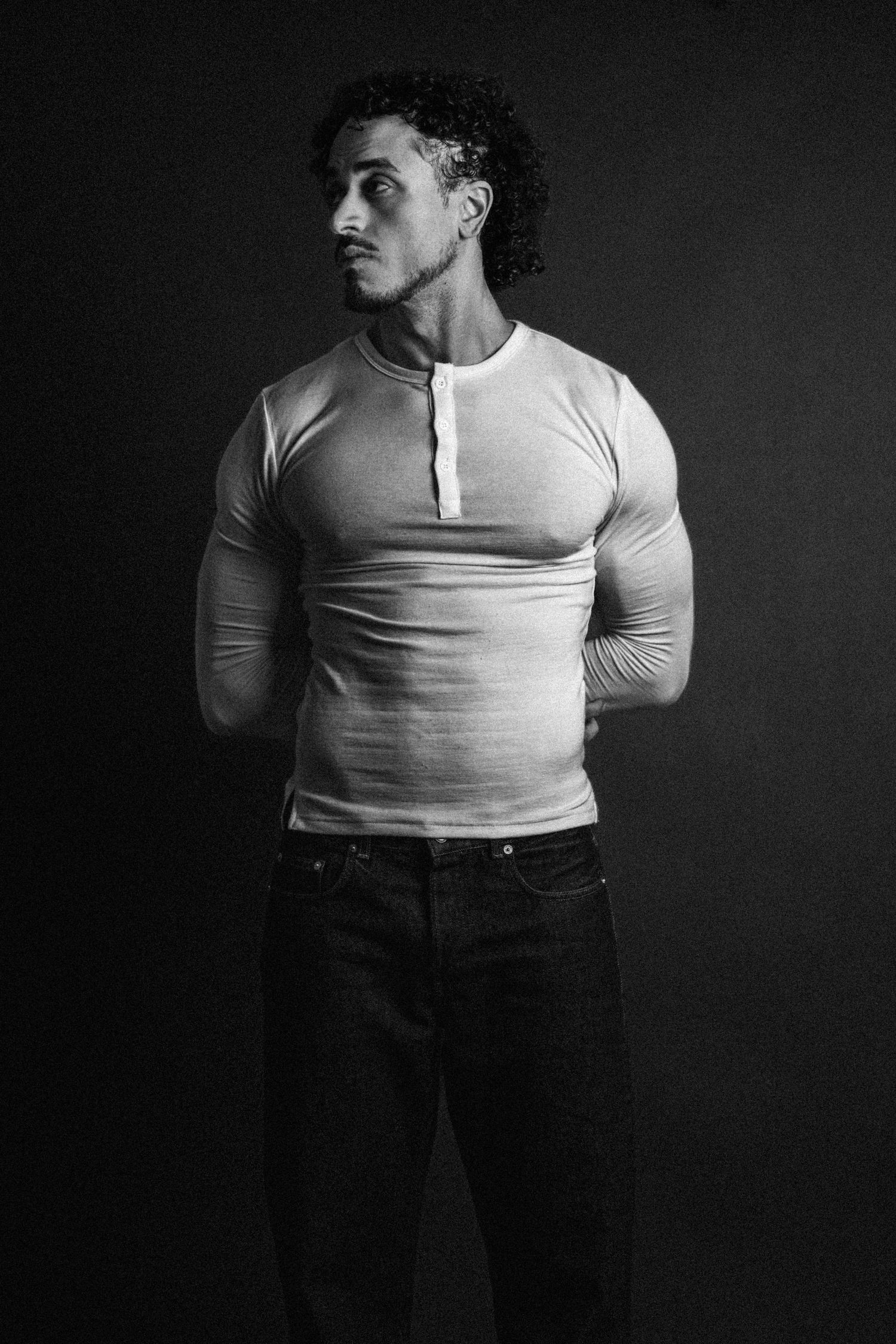 Black and white studio portrait of a muscular man with curly hair and beard in a fitted henley shirt and jeans, hands behind his back.