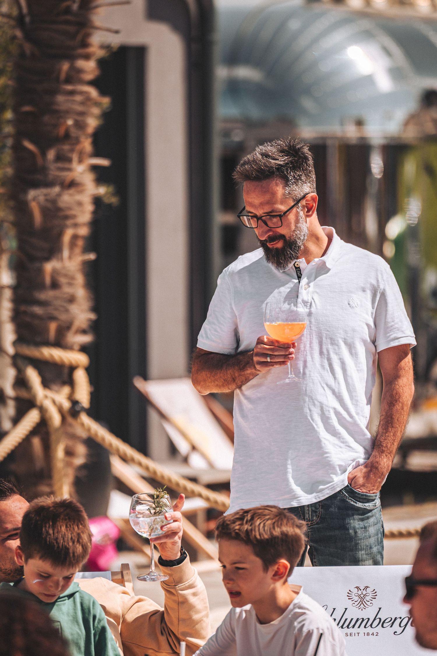Man in white polo shirt holding an orange cocktail at an outdoor beach bar restaurant event.