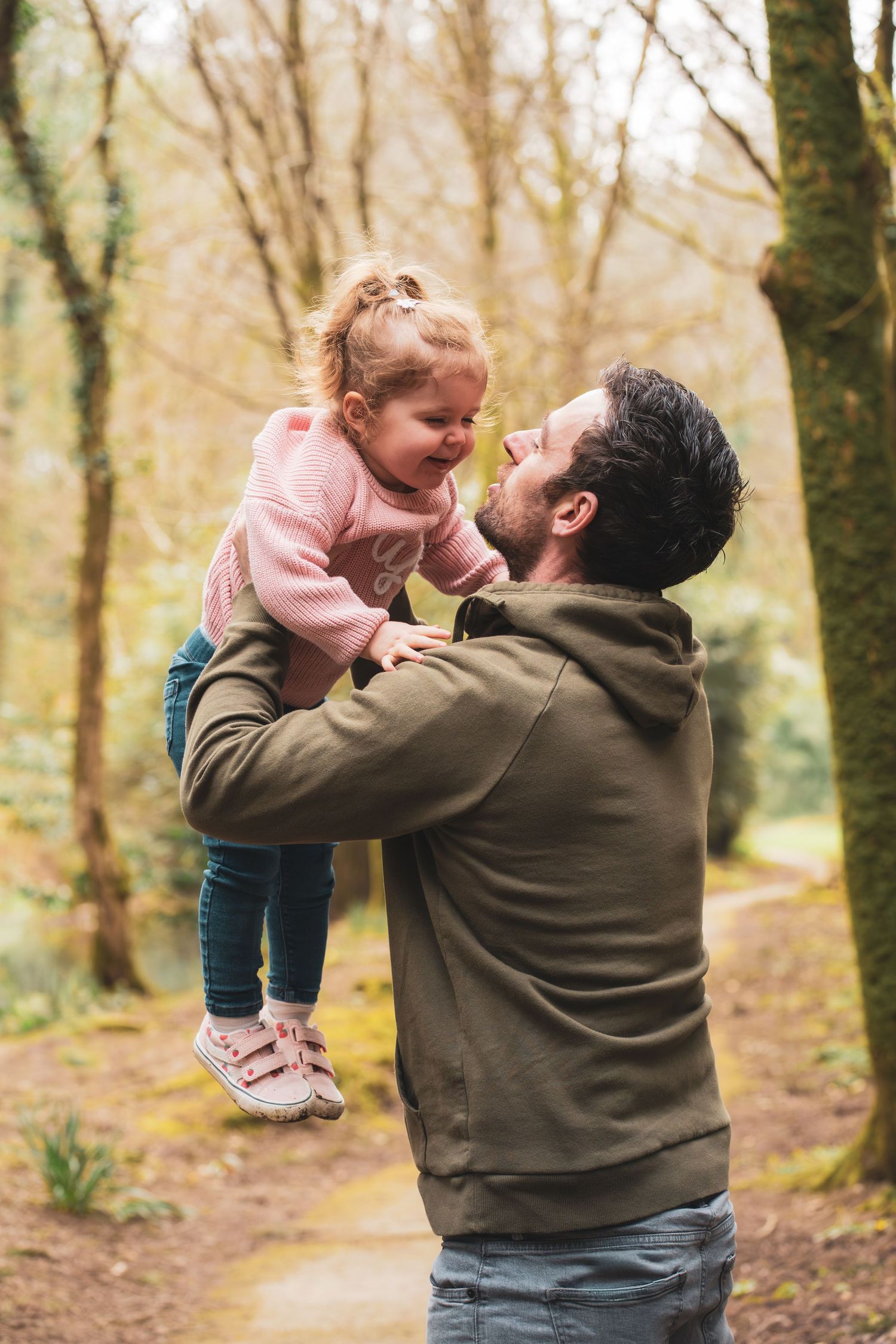An adult lifting a smiling young child during a walk in the woods.