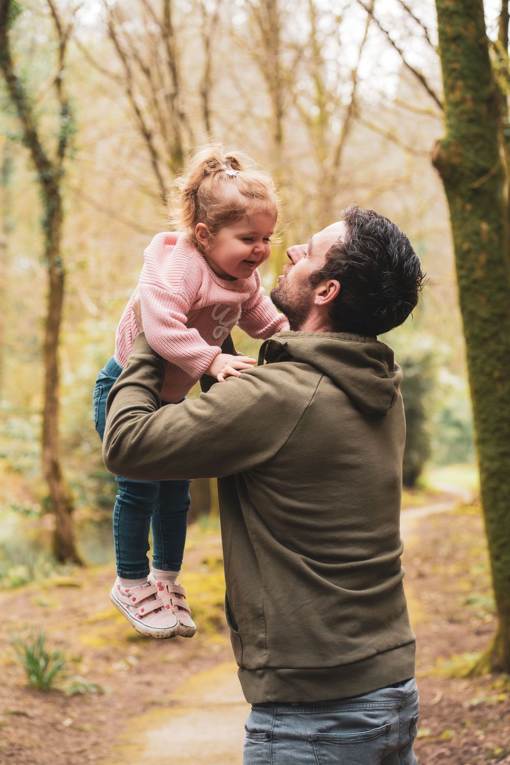 An adult lifting a smiling young child during a walk in the woods.