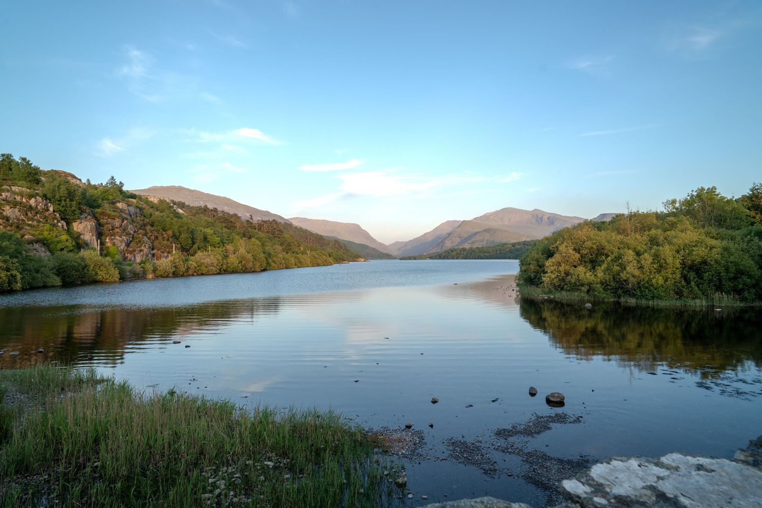Calm lake reflecting mountains and trees under a clear blue sky at dusk.