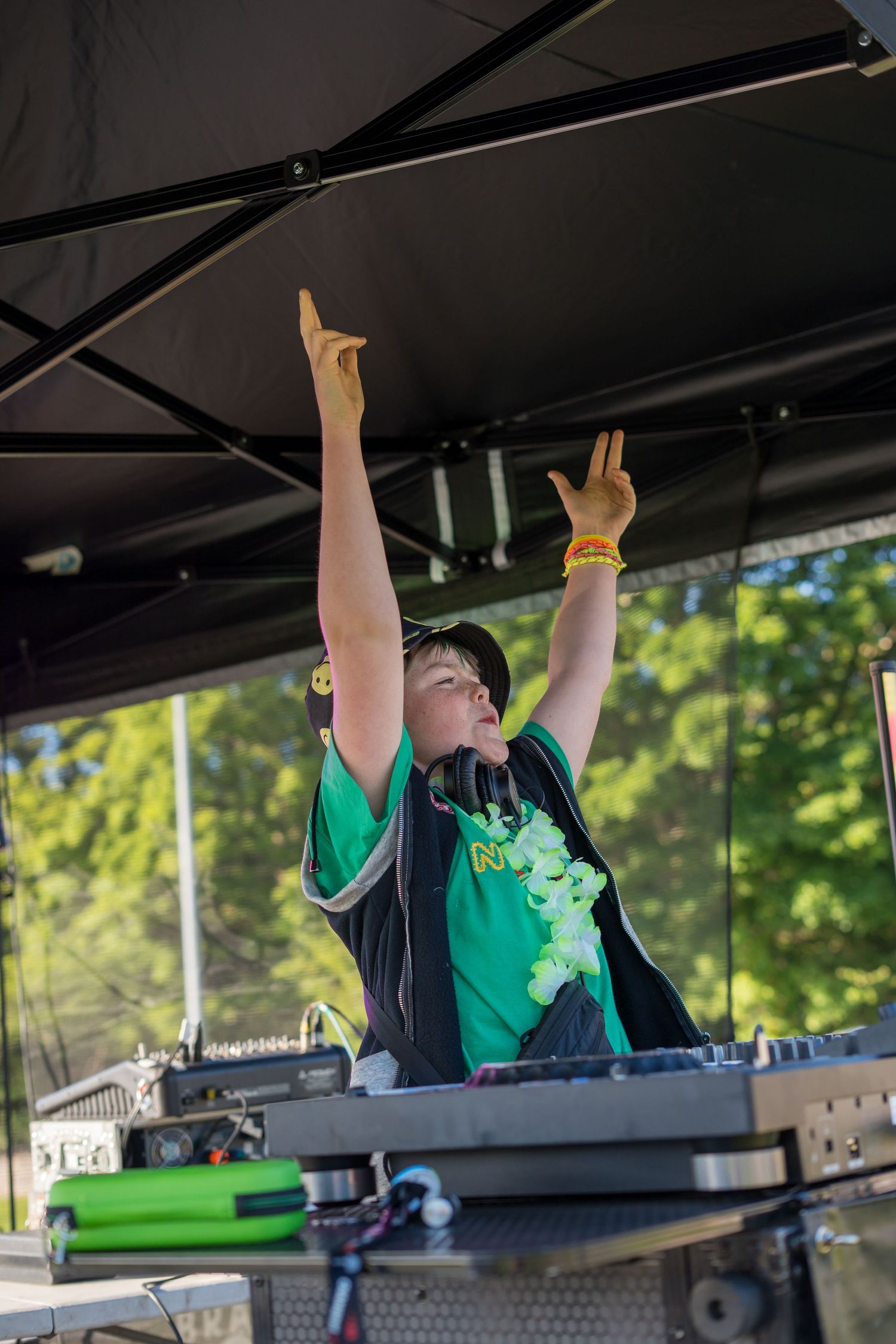 A DJ raising both arms behind mixing equipment on an outdoor stage.