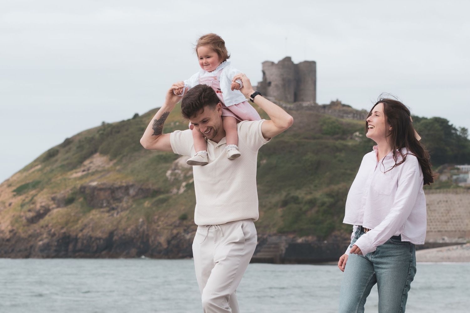 A happy family with a child on dad's shoulders enjoying a walk on a beach with a historic castle backdrop.