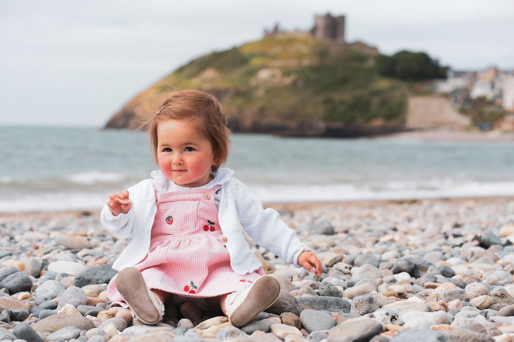 A young child sitting on a pebble beach with the sea and a castle-topped headland behind.