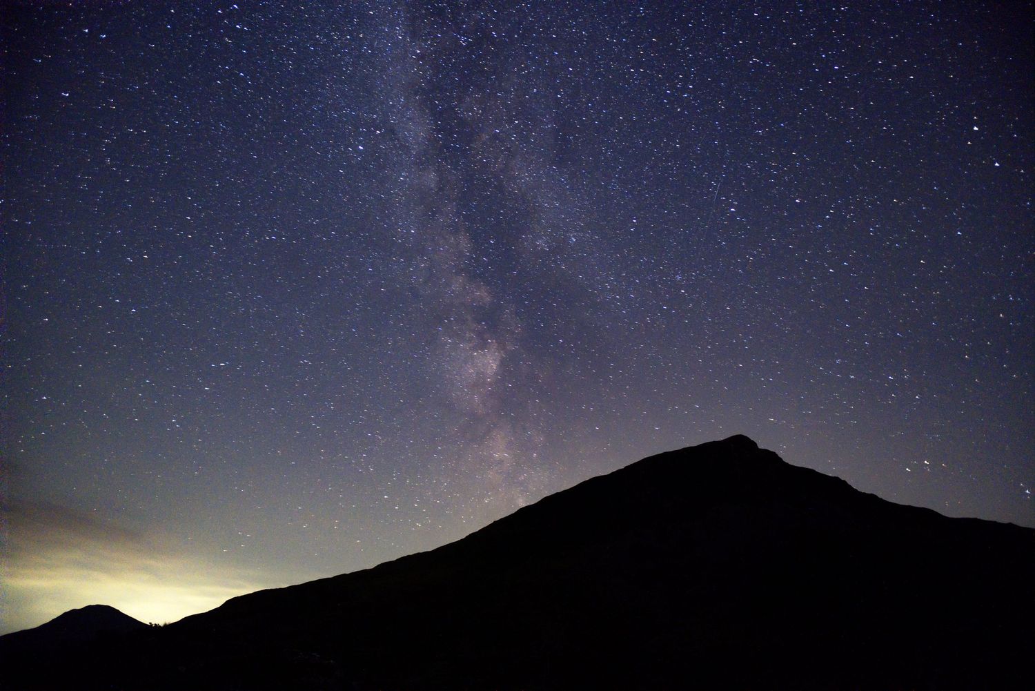Milky Way galaxy rising over a dark mountain silhouette in a starry night sky.