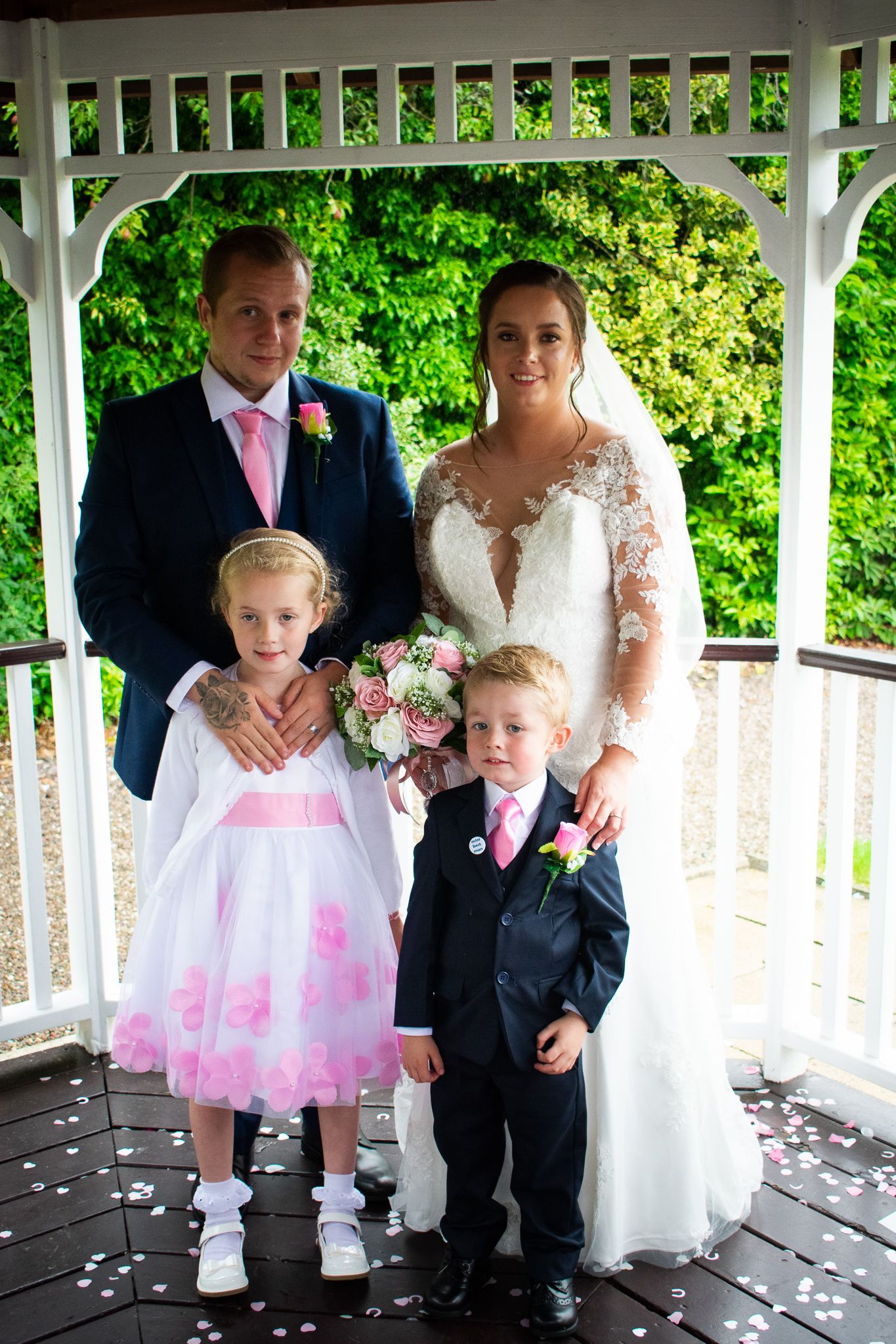 A wedding couple posing with two young children beneath a garden gazebo.