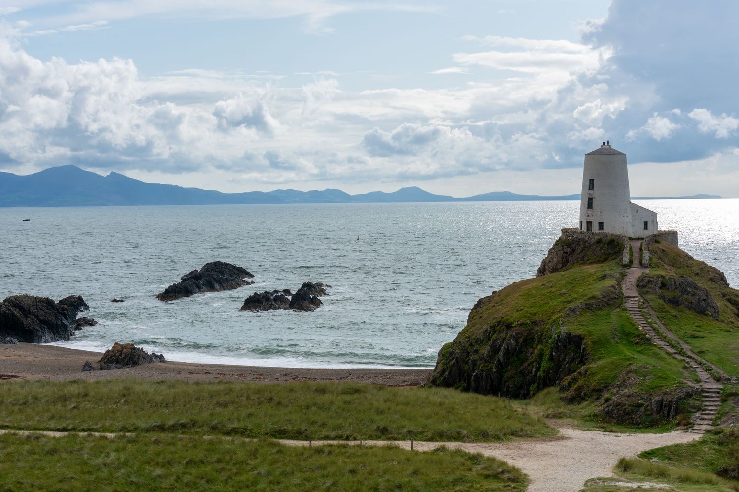 A stone tower on a grassy coastal headland overlooking the sea and distant mountains.