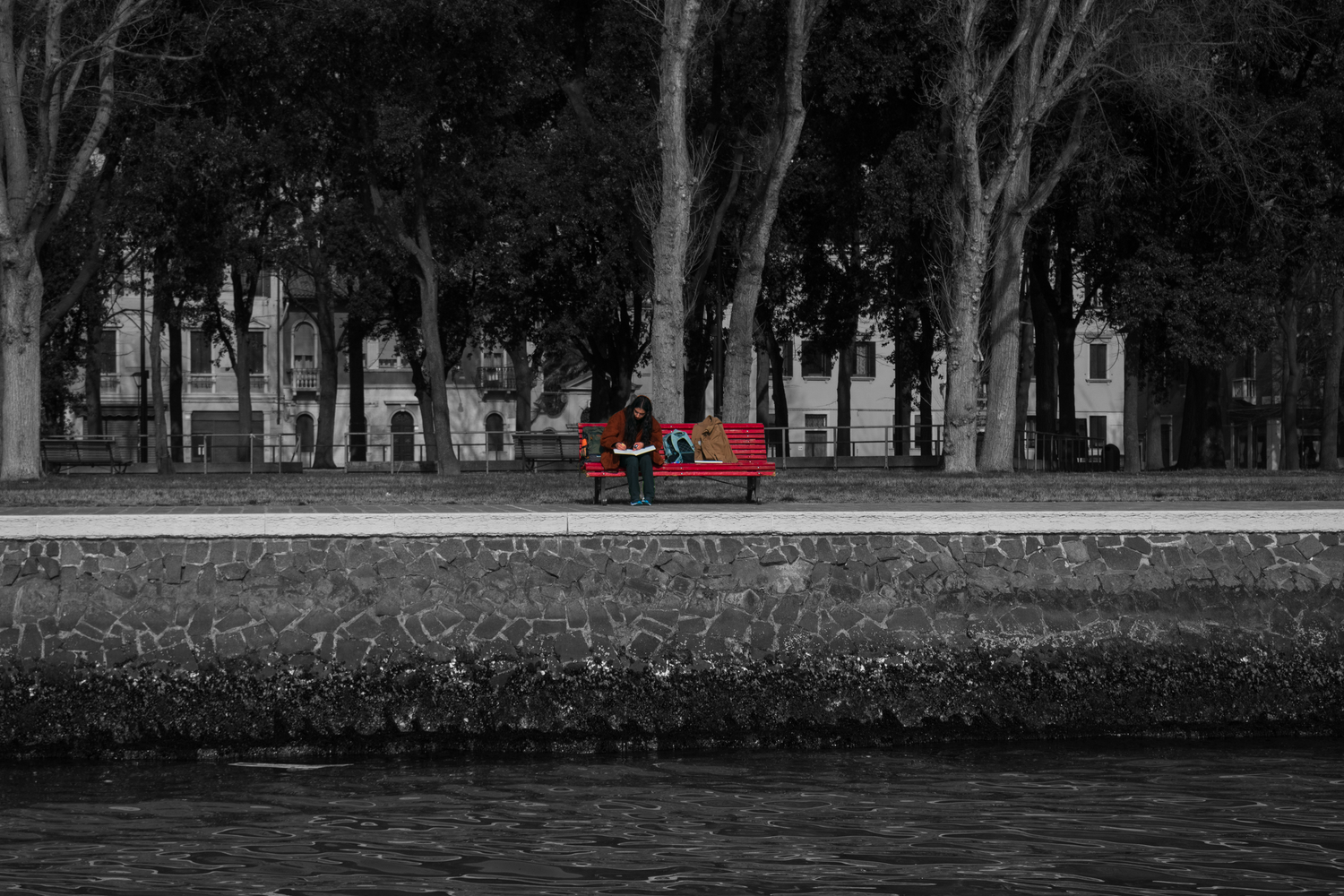 A red bench on a tree-lined path beside the water in an otherwise black and white photograph.