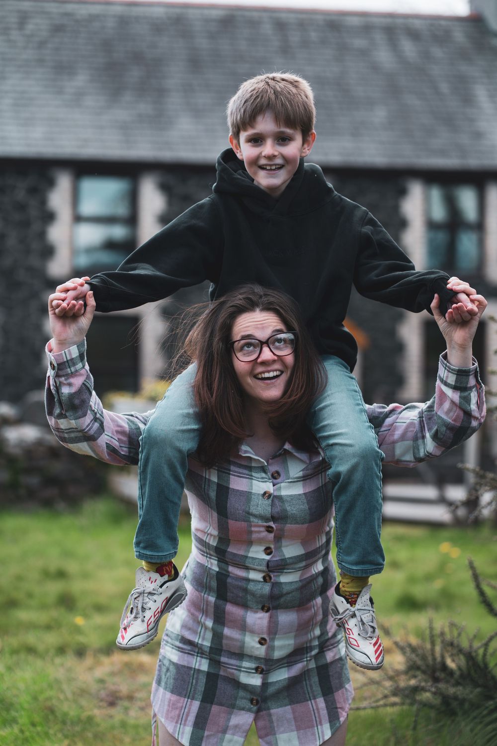 A smiling adult carrying a child on their shoulders in a garden.