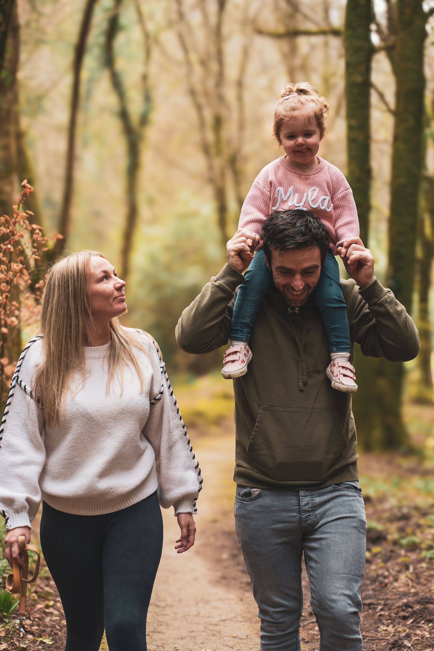 Two adults walking through woodland with a young child riding on one adult's shoulders.