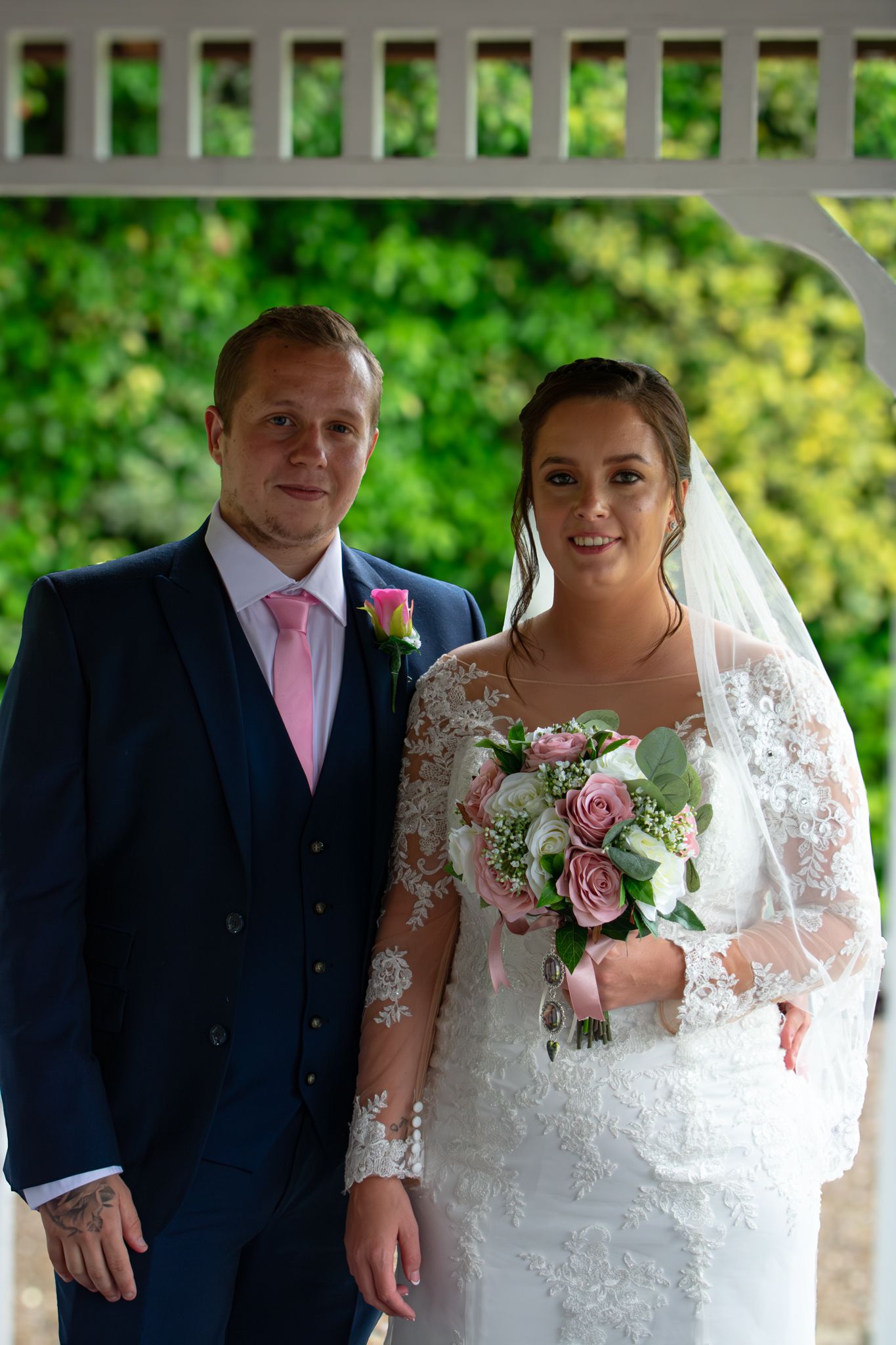 A wedding couple posing beneath a garden gazebo, with the bride holding a bouquet.