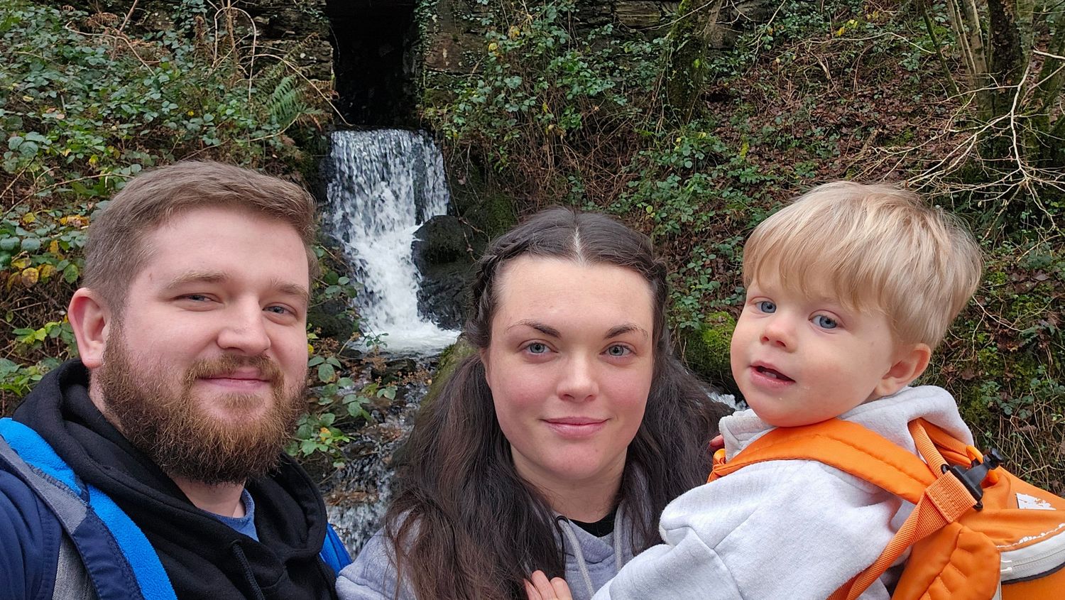 Two adults and a young child taking a selfie beside a woodland waterfall.