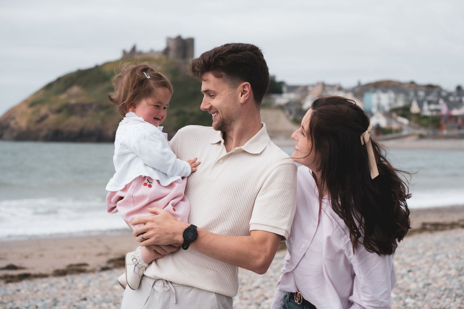 Two adults with a young child on a pebble beach, with a coastal castle in the background.