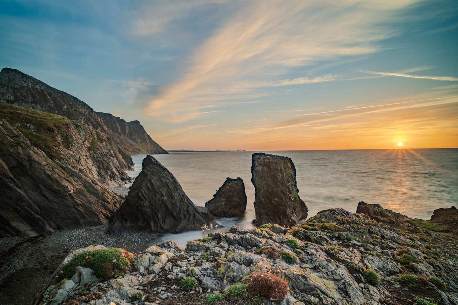 Dramatic coastal sunset with rugged sea stacks and cliffs silhouetted against a golden sky over calm ocean waters.