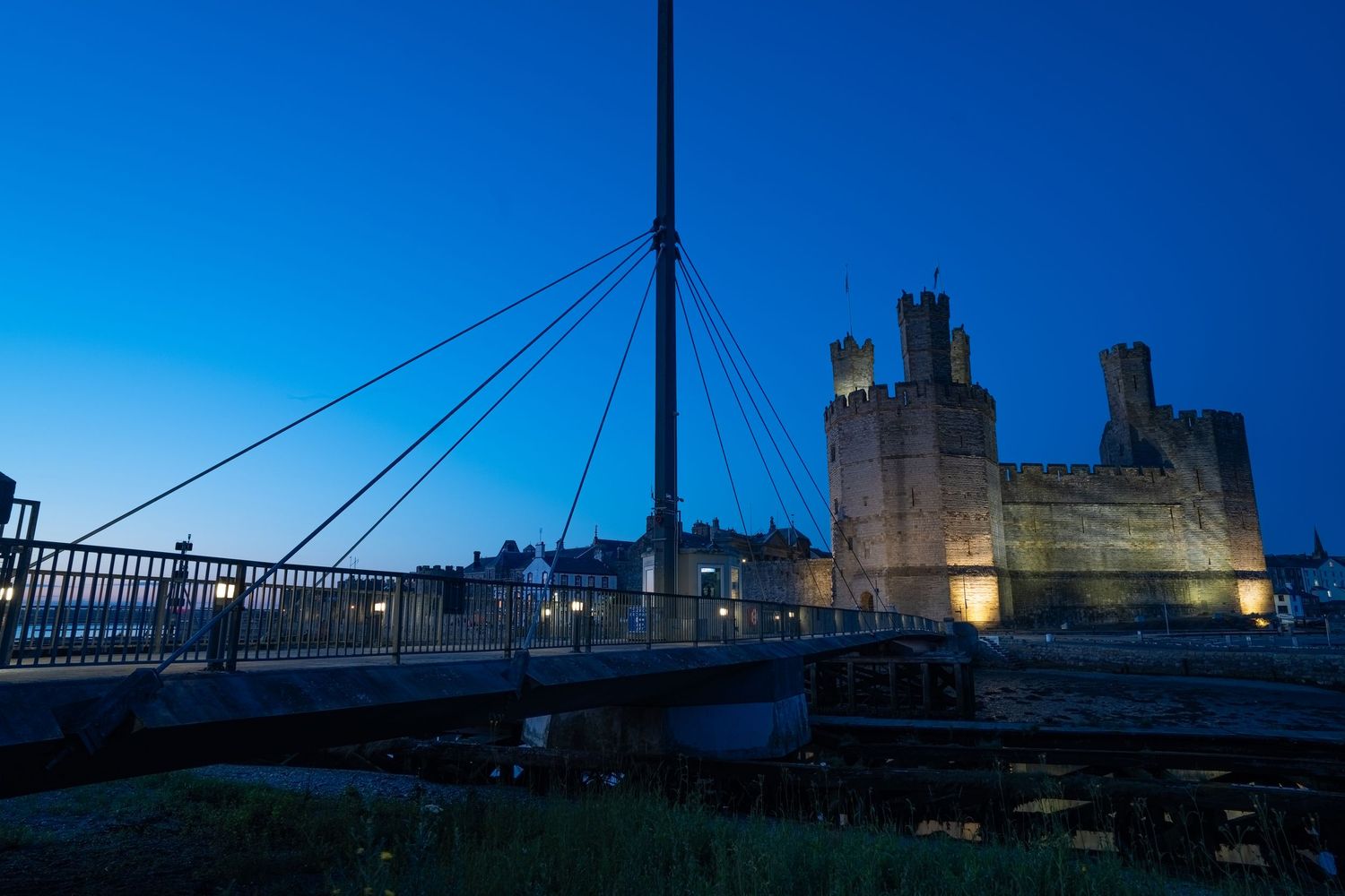 Caernarfon Castle illuminated at night with a modern suspension bridge in the foreground under blue sky.