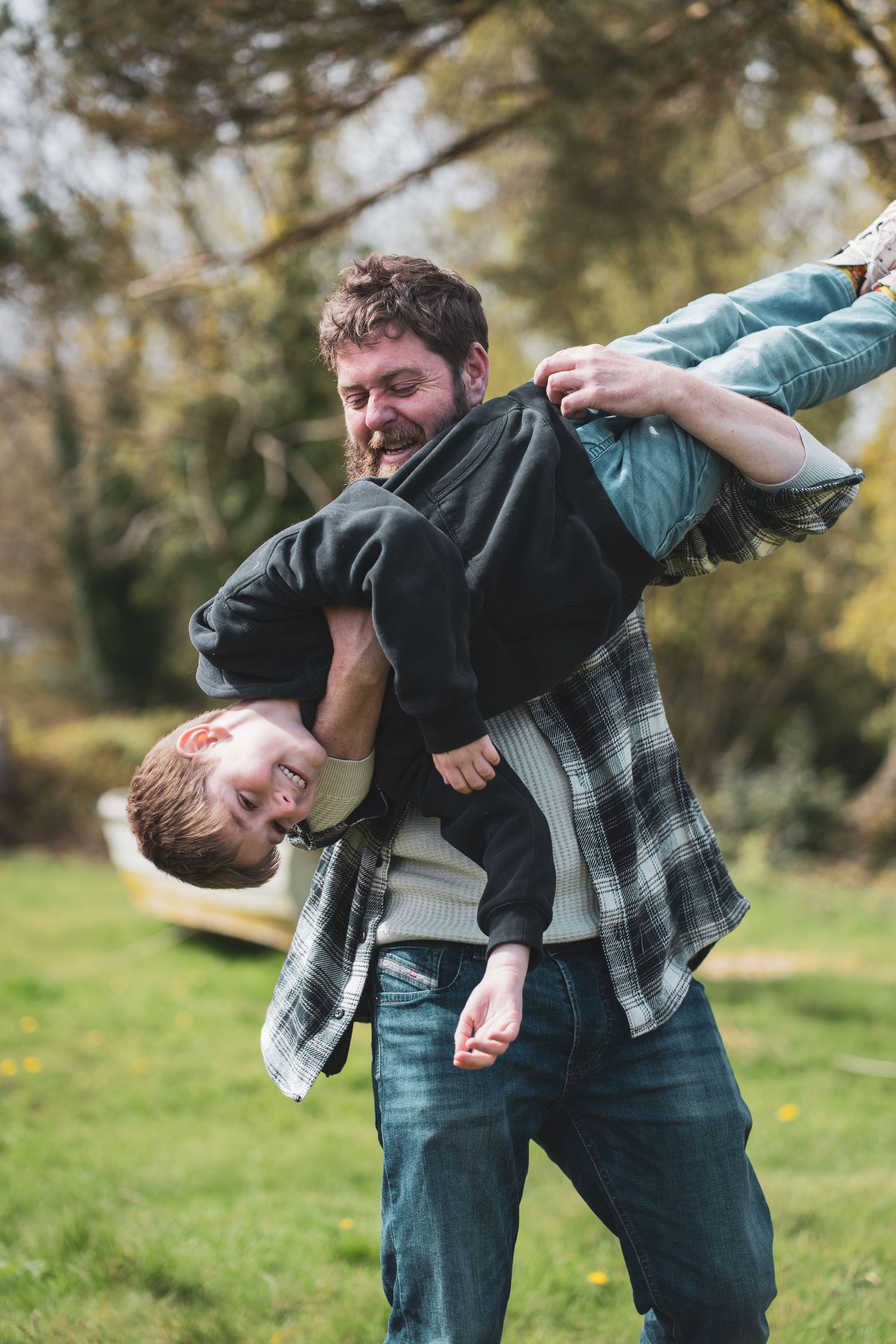 A smiling adult playfully carrying a child sideways over one shoulder outdoors.