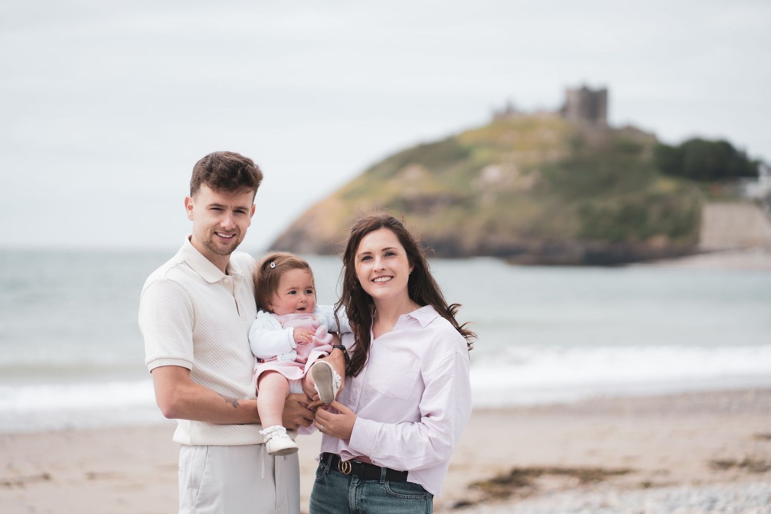 Two adults posing with a young child on a pebble beach beneath a castle-topped headland.
