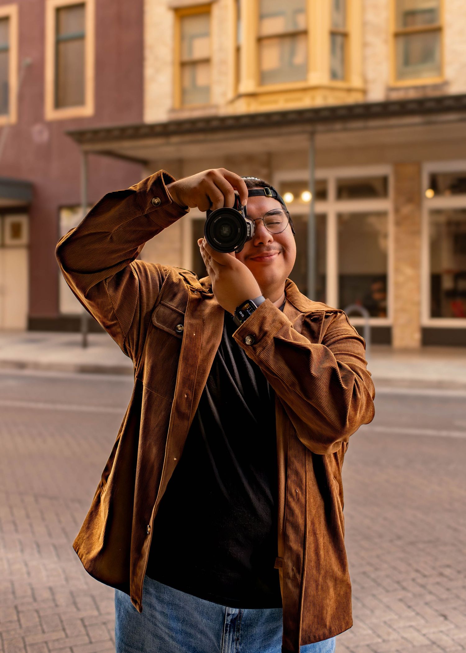 Smiling photographer in brown corduroy jacket holding camera on urban street.