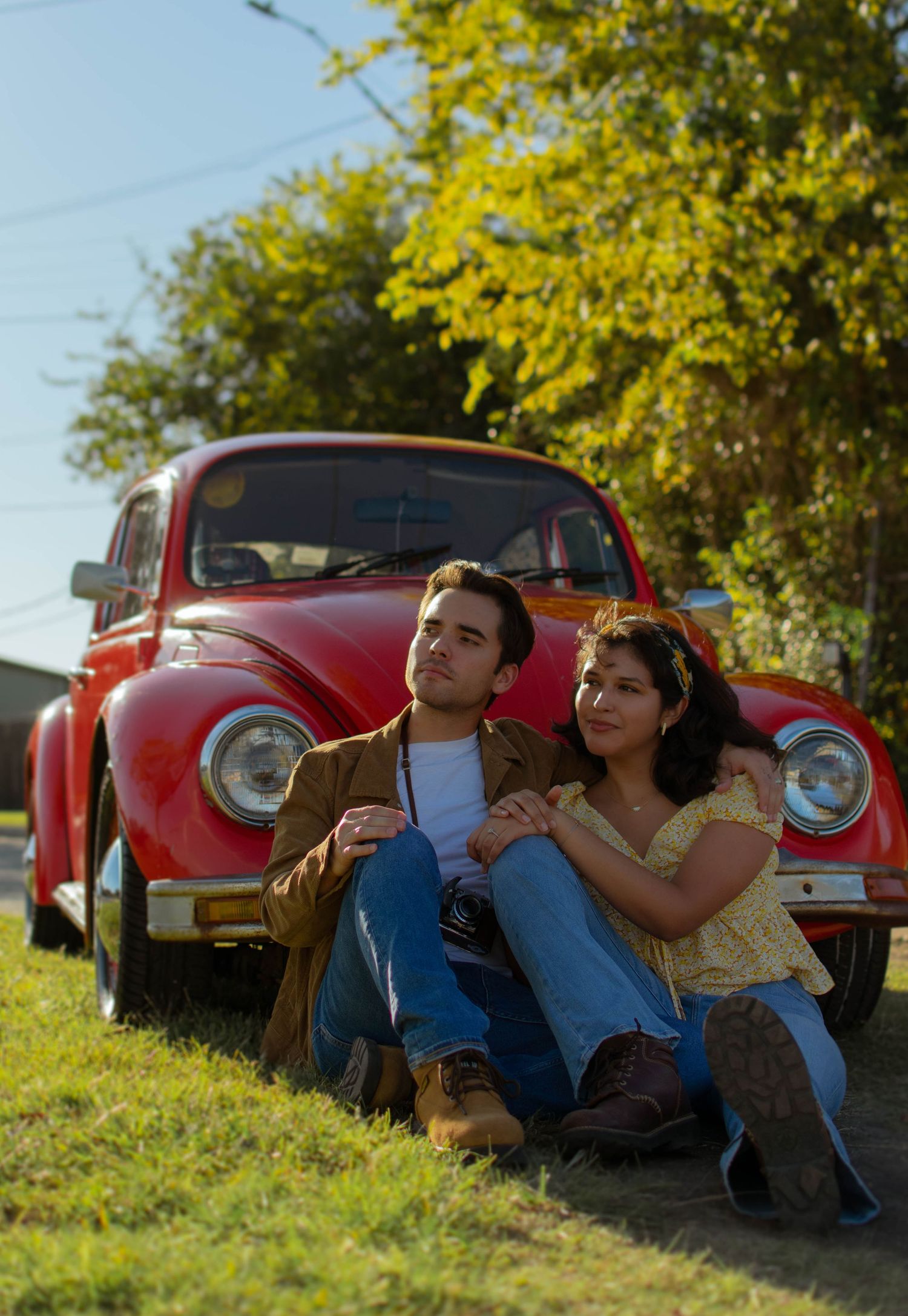 A young couple sits together on grass leaning against a classic red Volkswagen Beetle outdoors.