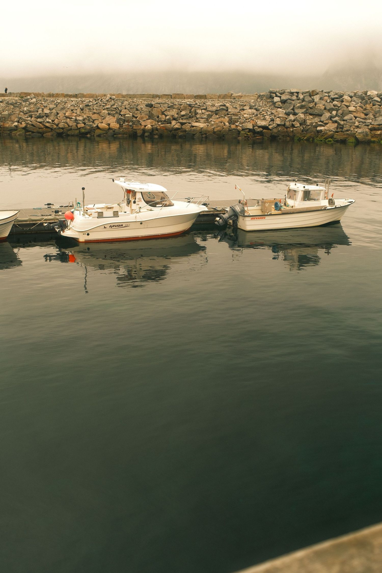 Two fishing boats moored in a calm, foggy harbour behind a stone breakwater on Senja, Norway