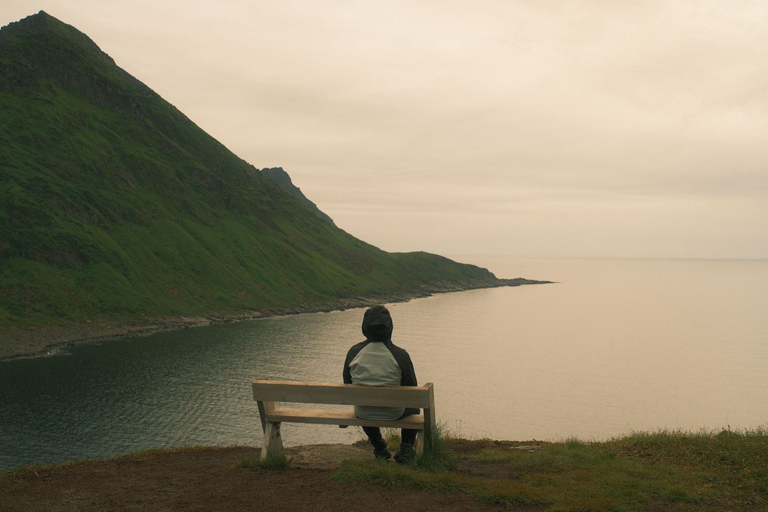 Man sitting on a wooden bench above a calm fjord beneath a green mountain on Senja, Norway
