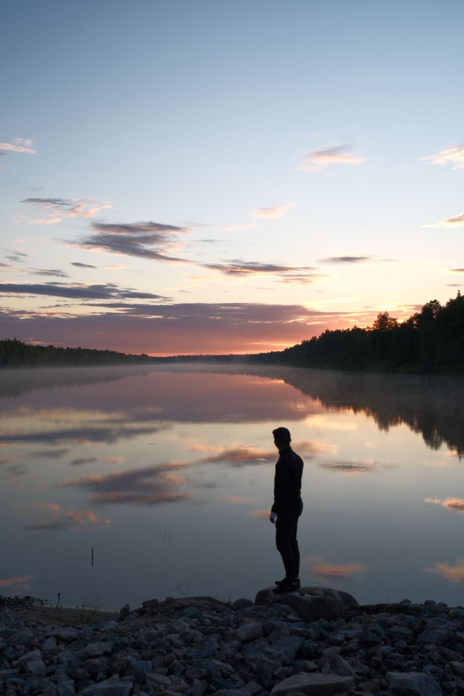 Silhouetted figure beside a mirror-calm lake under a pink Nordic summer sky