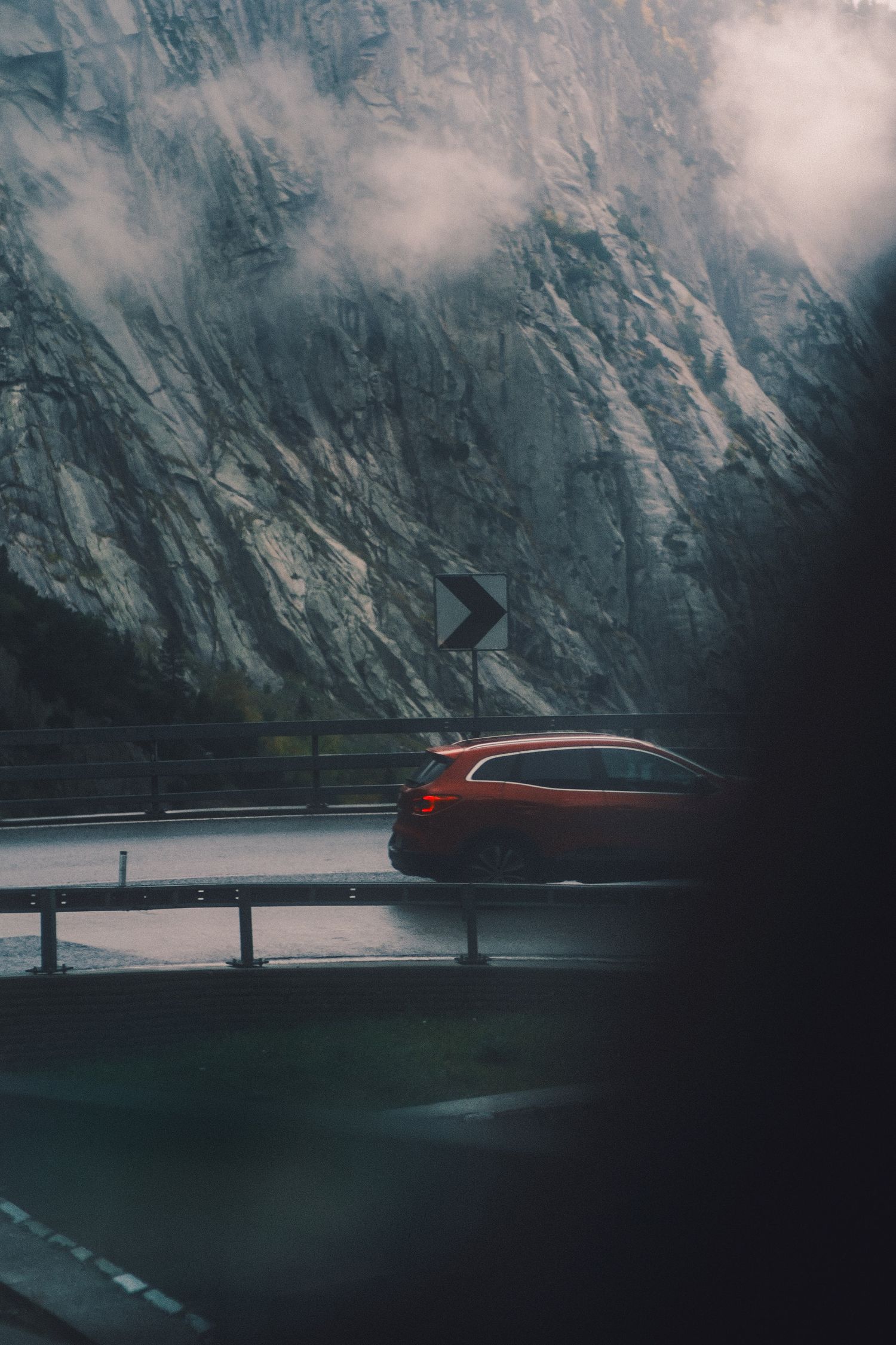 Red car on a wet mountain road beneath a sheer rock face on the Gotthard Pass, Switzerland