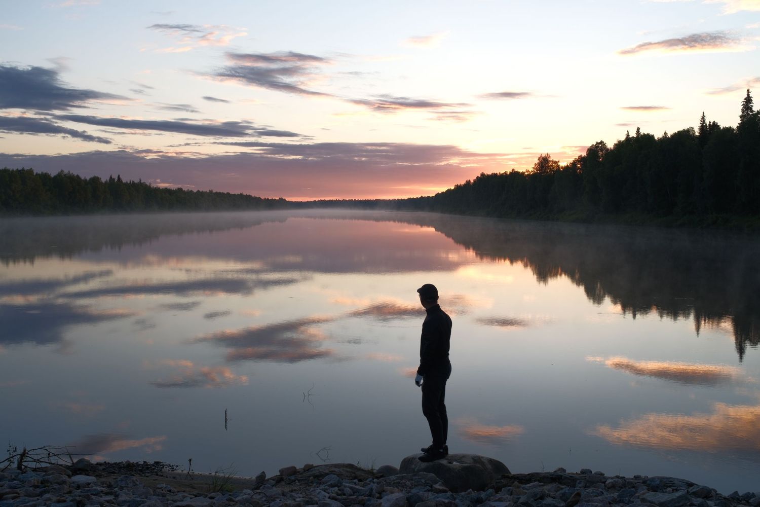 Lone figure at the edge of a still Scandinavian lake in the midnight sun