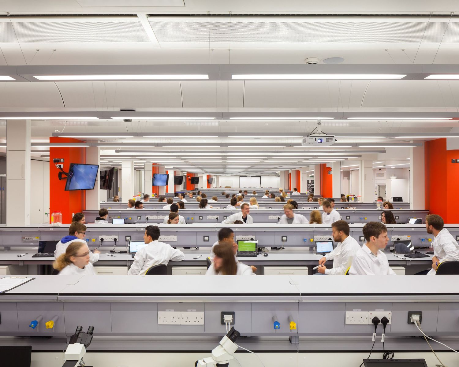 Students in white lab coats work at modern laboratory benches in a large, bright science classroom with orange accent walls.