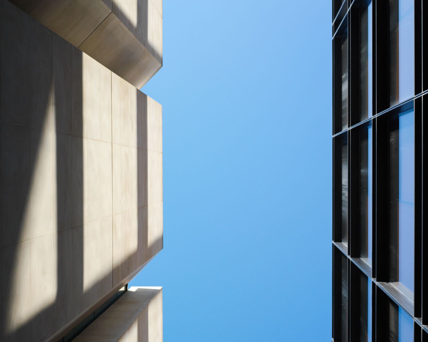 Two modern buildings frame a clear blue sky, with concrete architecture and glass windows creating geometric contrast.