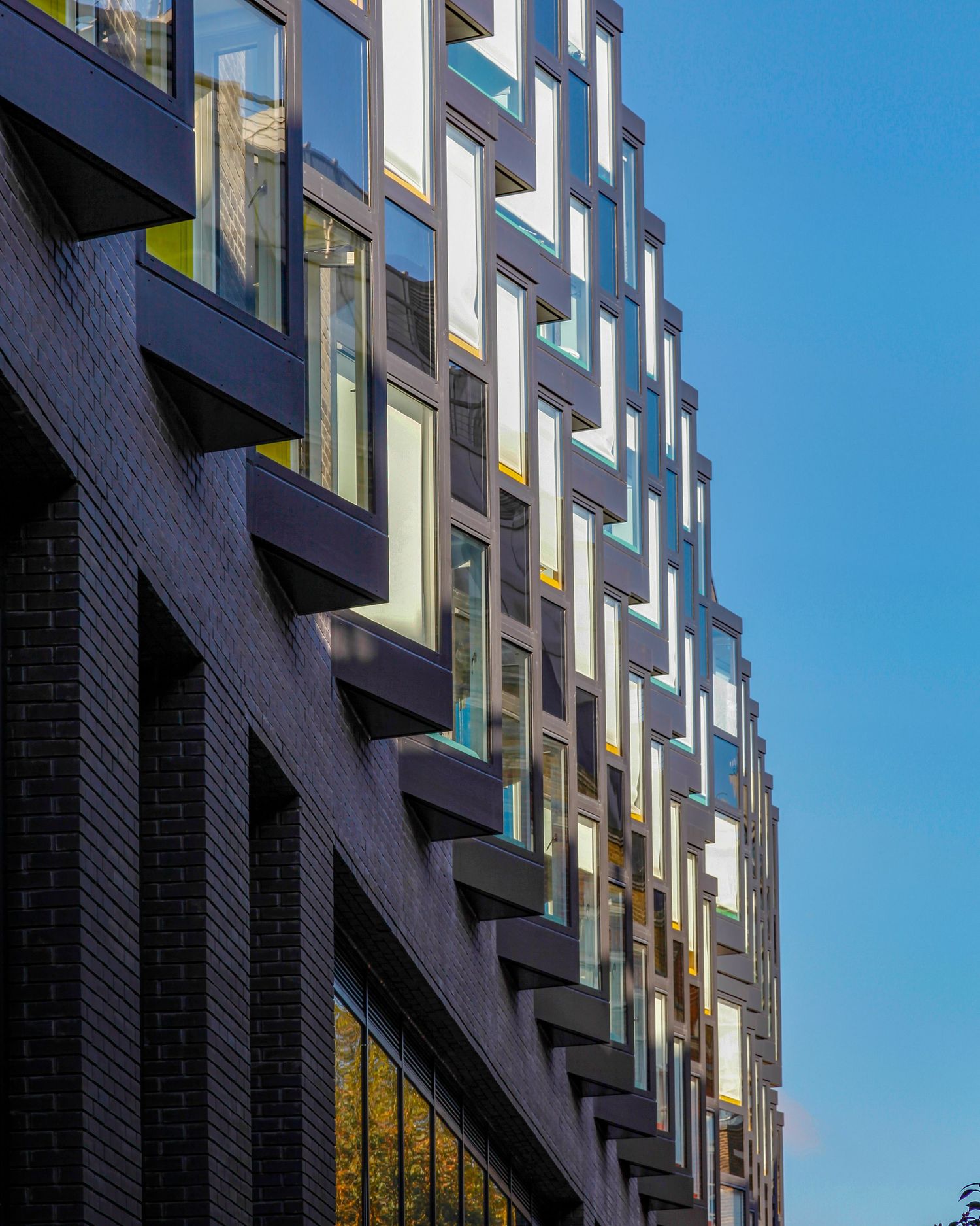 Modern dark brick building facade with geometric angular windows reflecting blue sky in contemporary urban architecture.