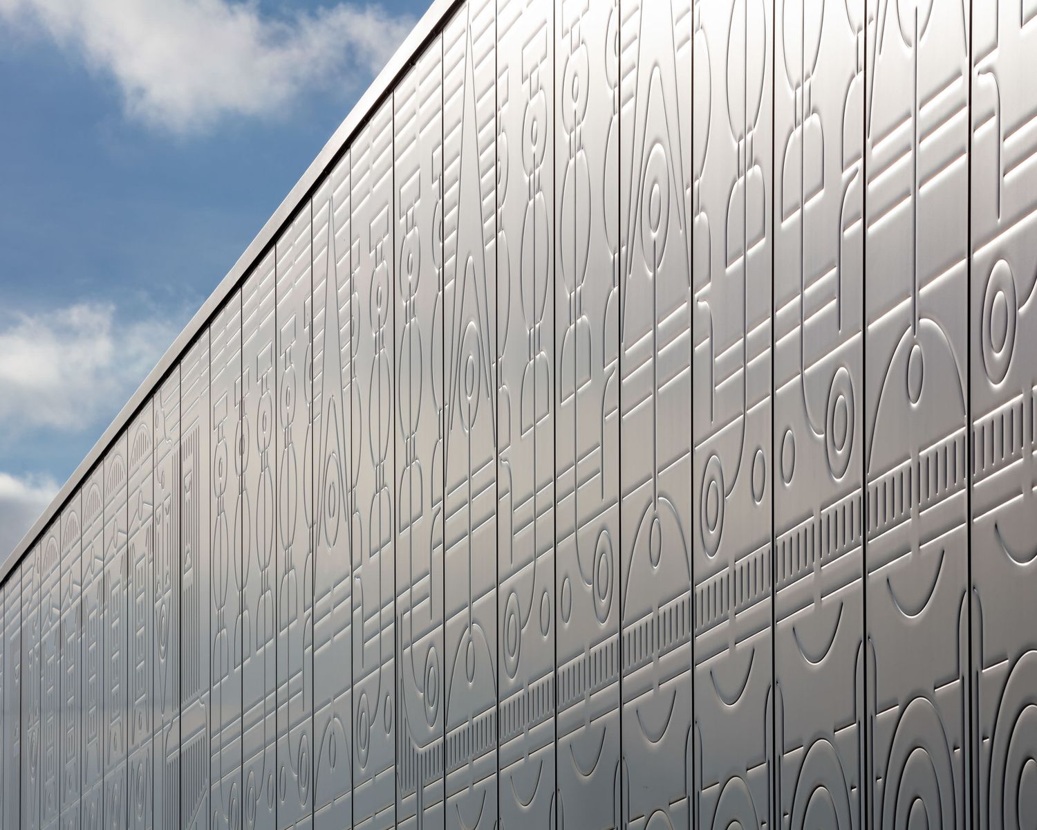 Modern building facade with decorative embossed silver metal panels featuring geometric patterns against a blue sky.