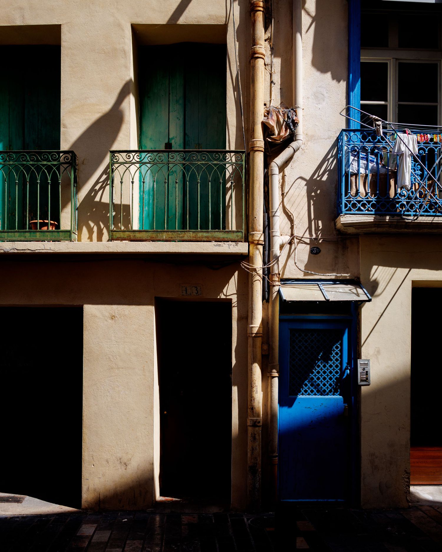 Sunlit building facade with green iron balconies, drainpipe, and vibrant blue door casting dramatic shadows.