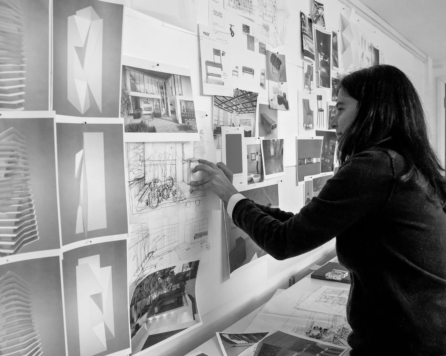 Black and white photo of a woman pinning architectural sketches and design mood board images to a studio wall.