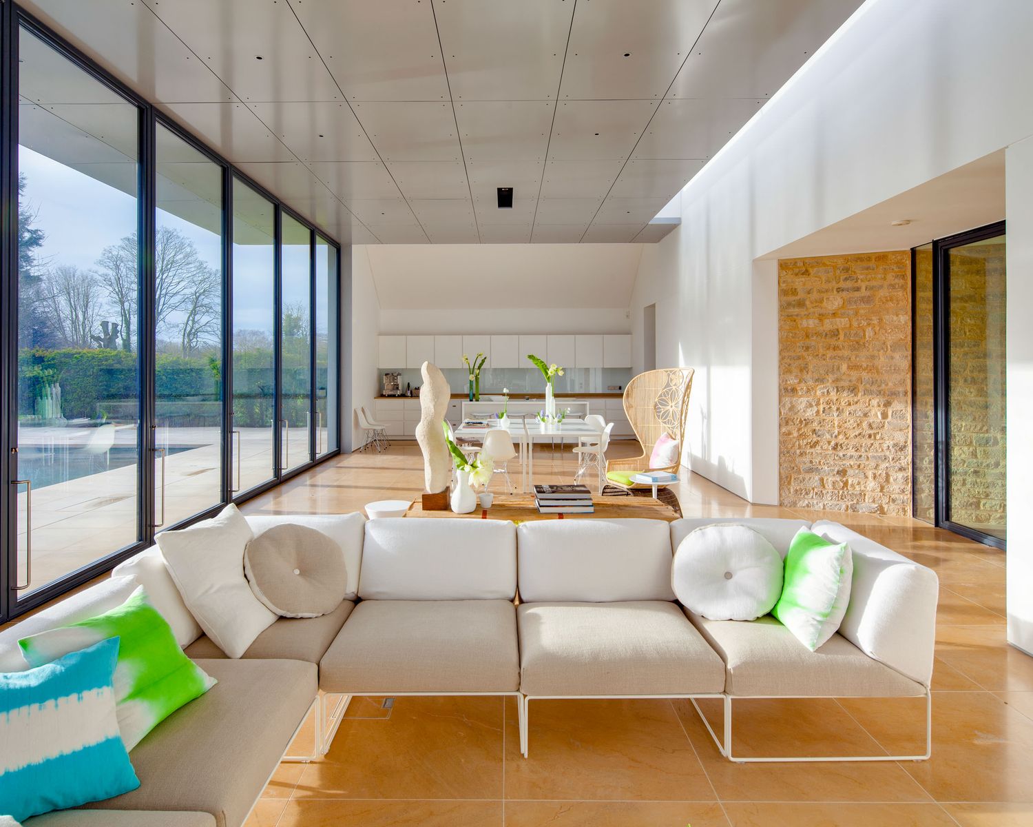 Modern open-plan living room with white sectional sofa, colorful cushions, floor-to-ceiling windows, and stone accent wall.
