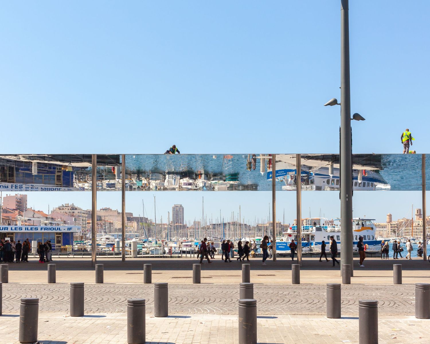 Mirrored canopy reflects harbor and pedestrians on a sunny waterfront promenade with bollards and clear blue sky.