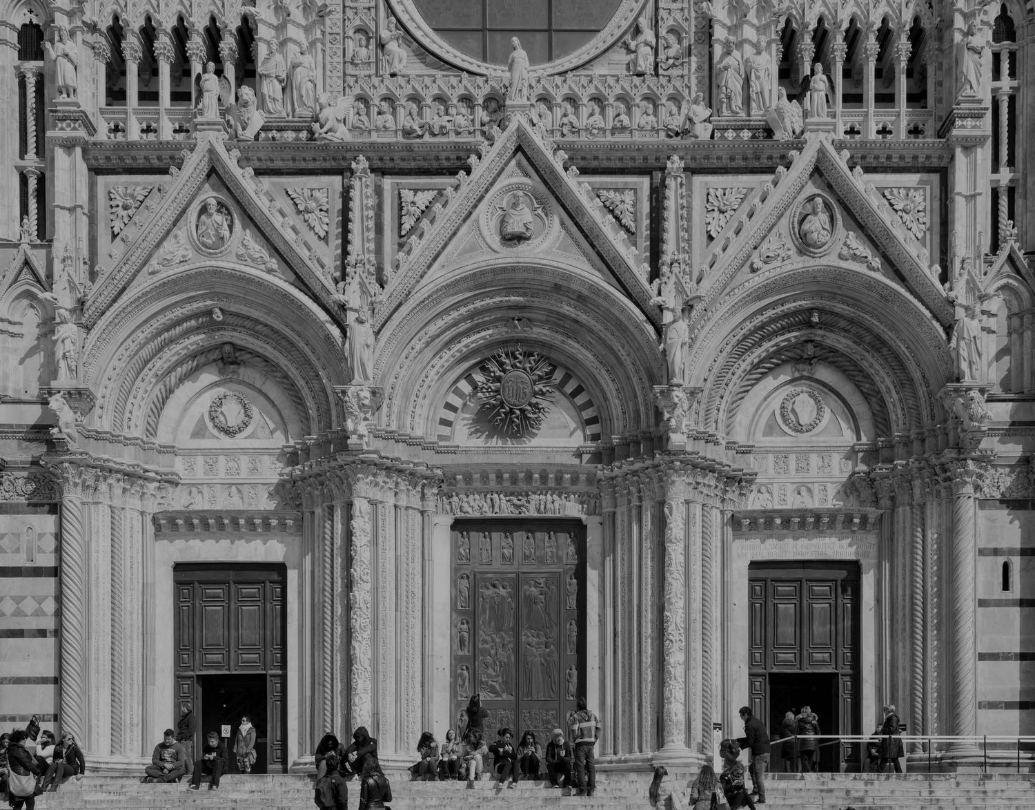 Black and white photo of the ornate Gothic facade of Siena Cathedral, Italy, featuring three arched portals and intricate carvings.