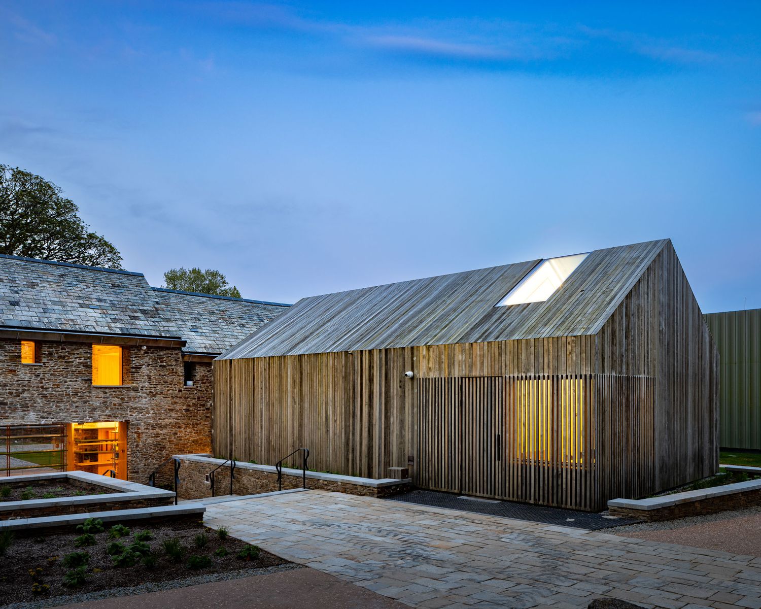 Modern barn conversion with timber cladding and zinc roof glows warmly at dusk beside a traditional stone farmhouse.