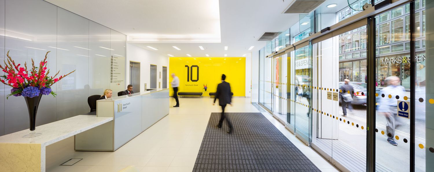 Modern building lobby with yellow accent wall, reception desk, and glass entrance doors.