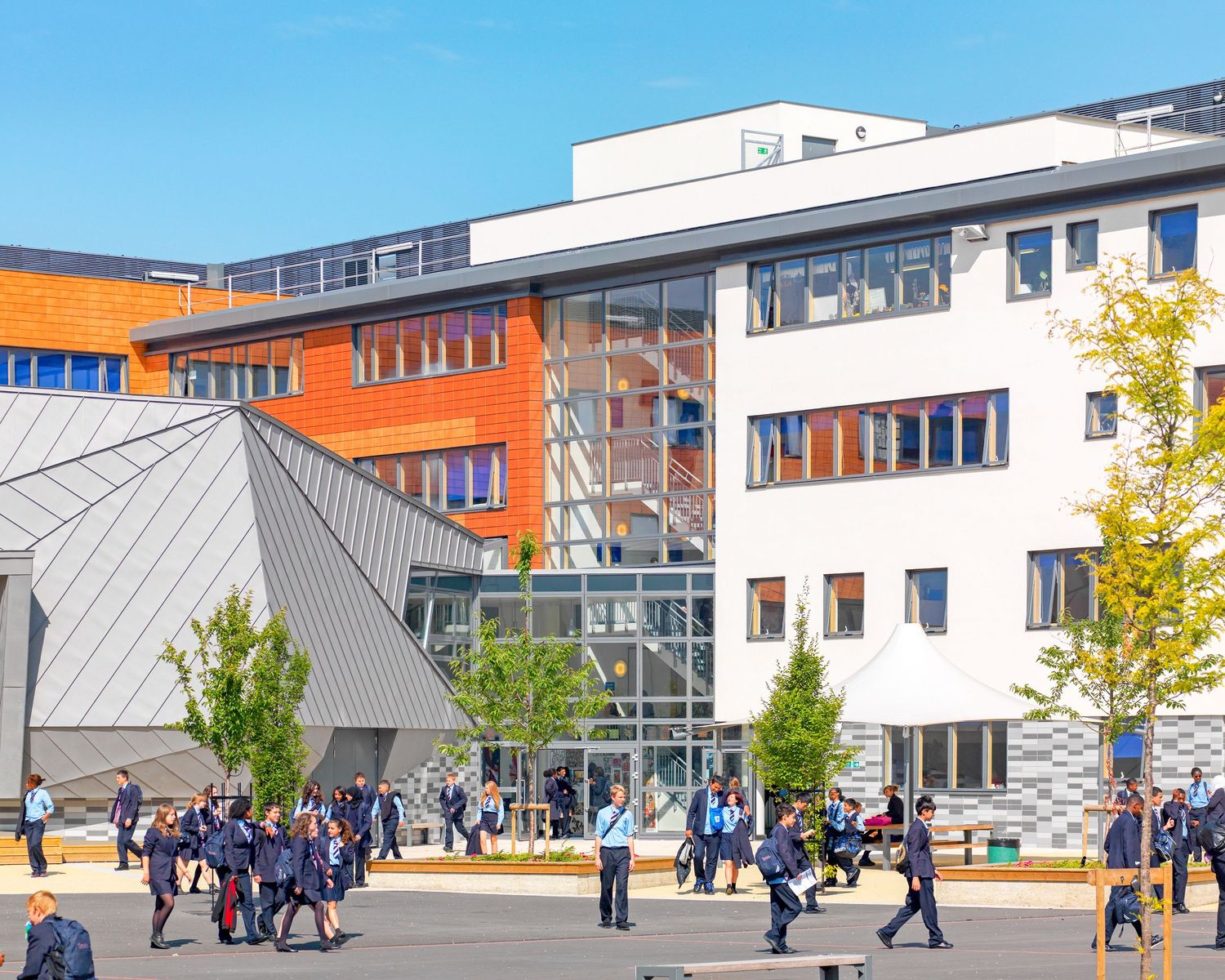 Students walking outside a modern school building with white facade, orange accents, and large glass windows on a sunny day.