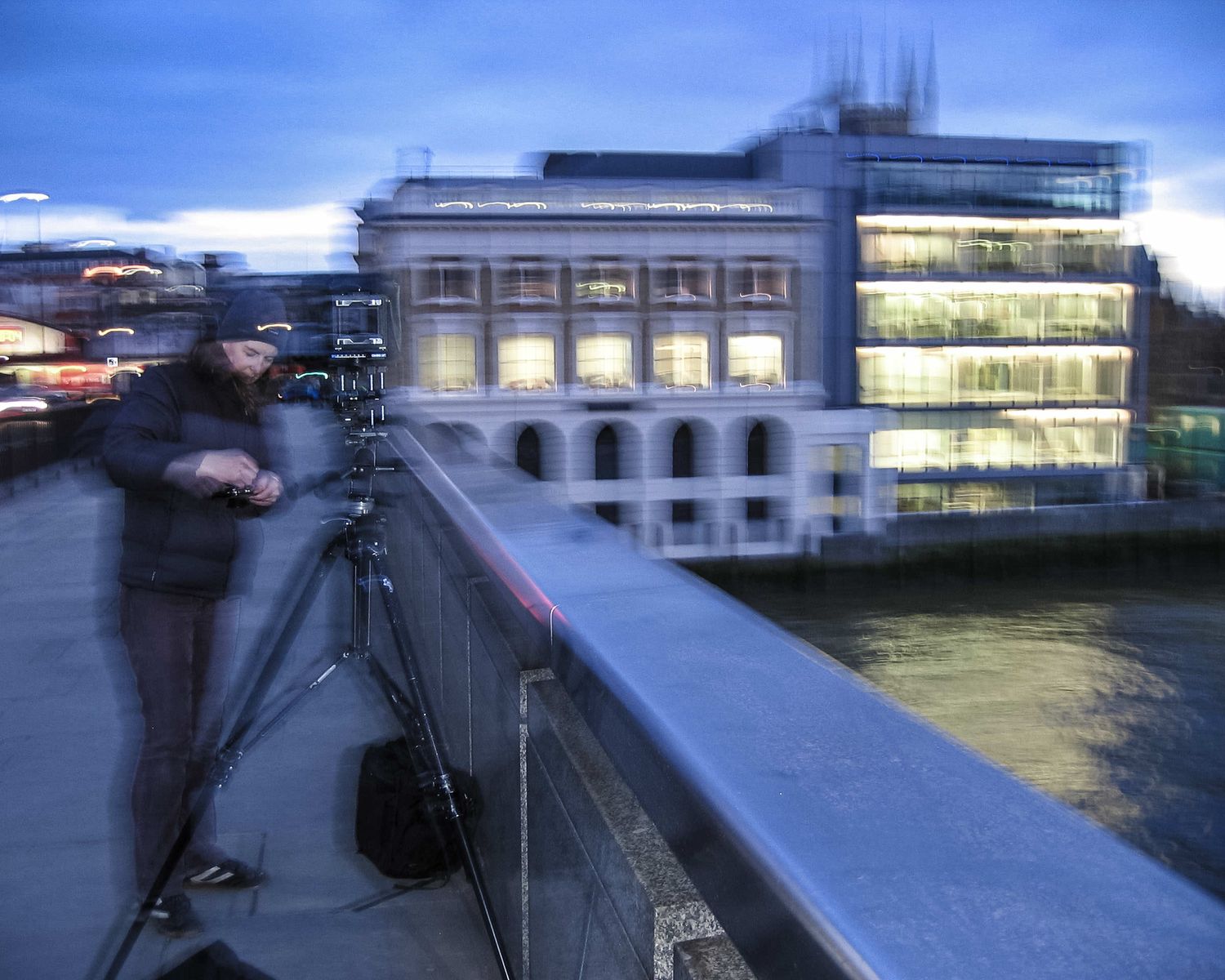 Photographer using a tripod on a bridge at dusk, with an illuminated classical building reflected on the river.