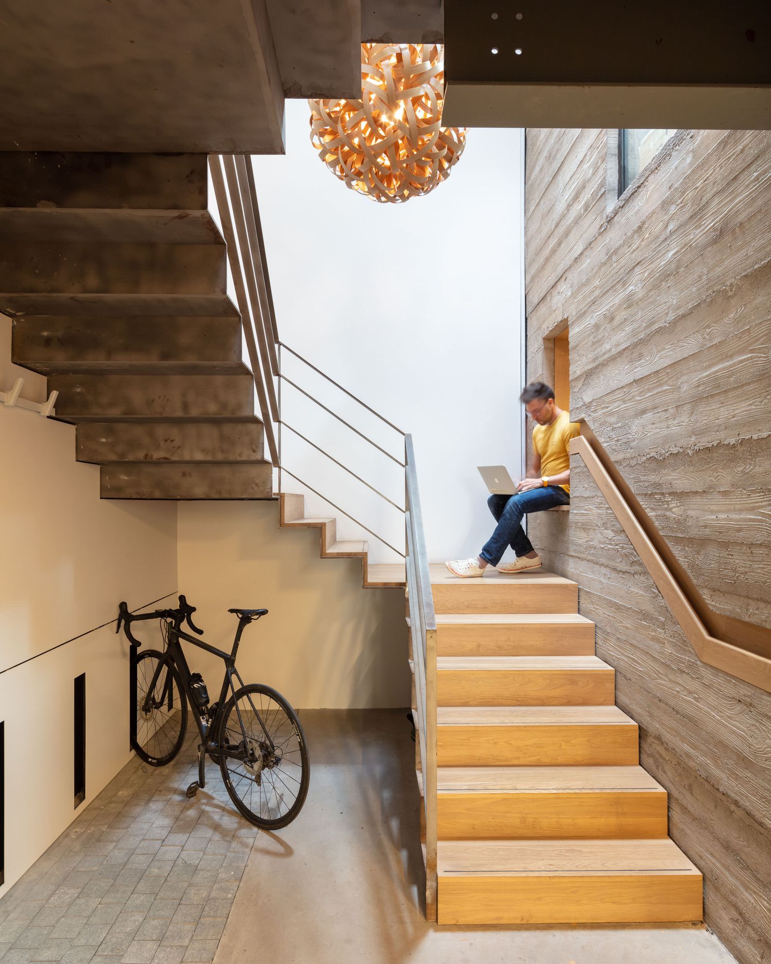 Person sitting on modern wooden staircase with laptop beside a bicycle and elegant chandelier.