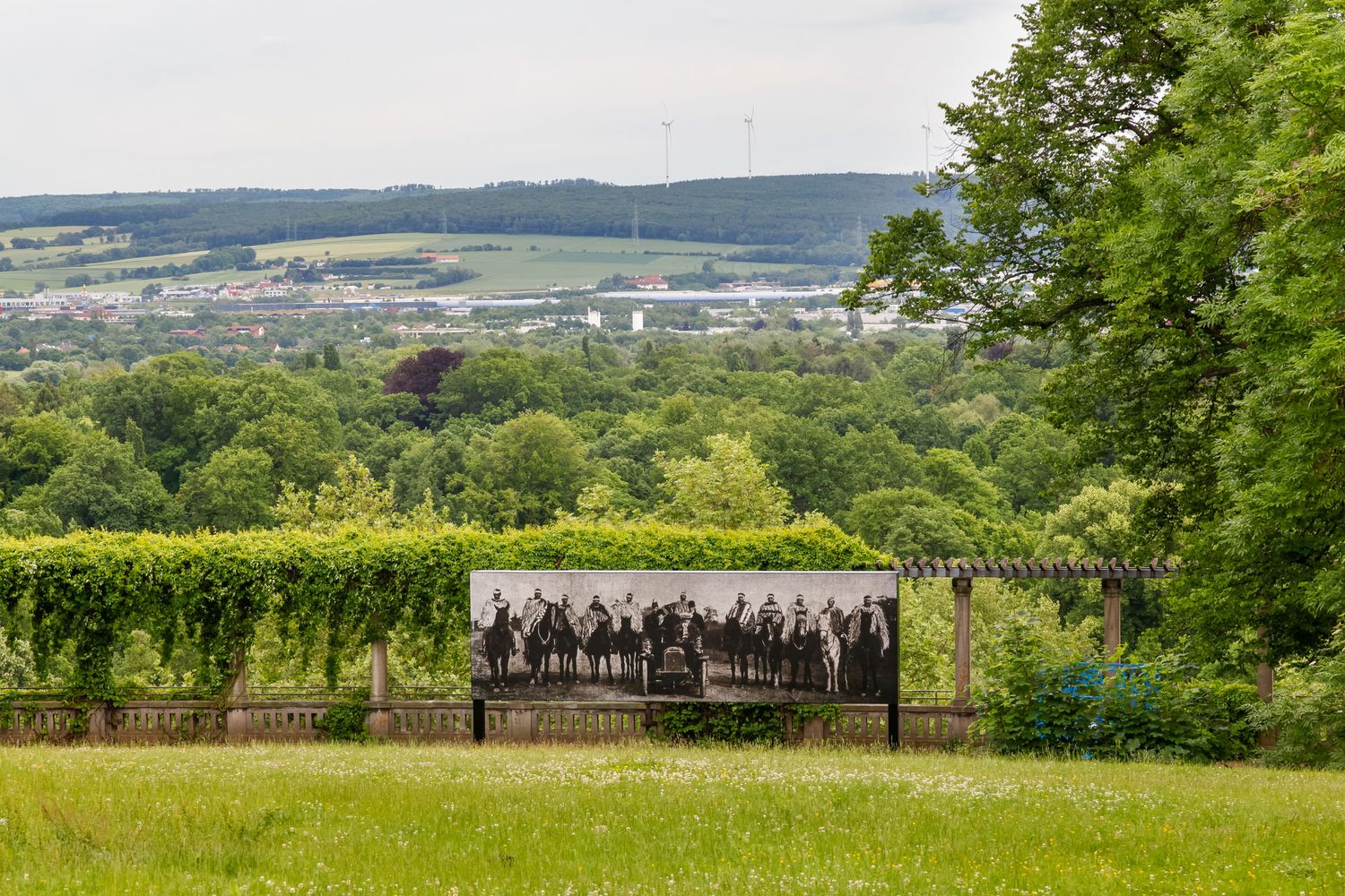 Scenic hilltop garden with a large historic group photograph display framed by lush green trees and rolling countryside.