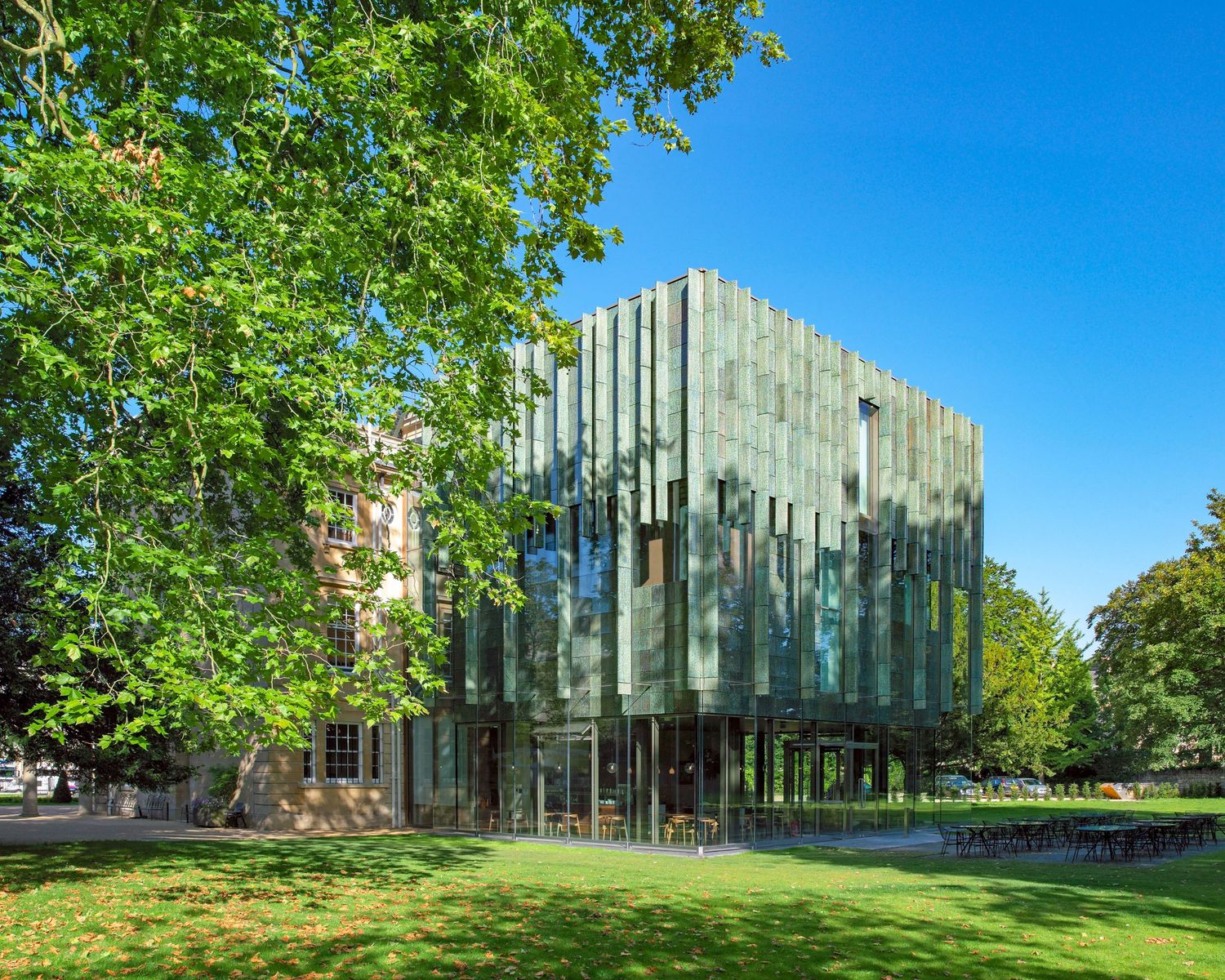 Modern glass building elevated on pillars surrounded by lush green trees under a clear blue sky.