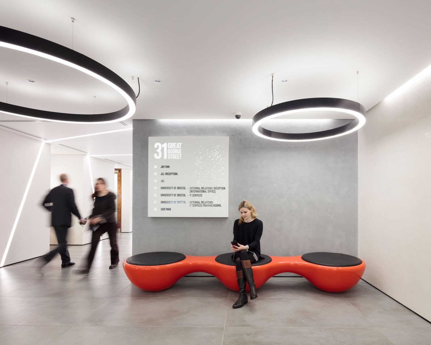 Modern office lobby with circular pendant lights, red sculptural seating, and blurred people walking past a directory wall.