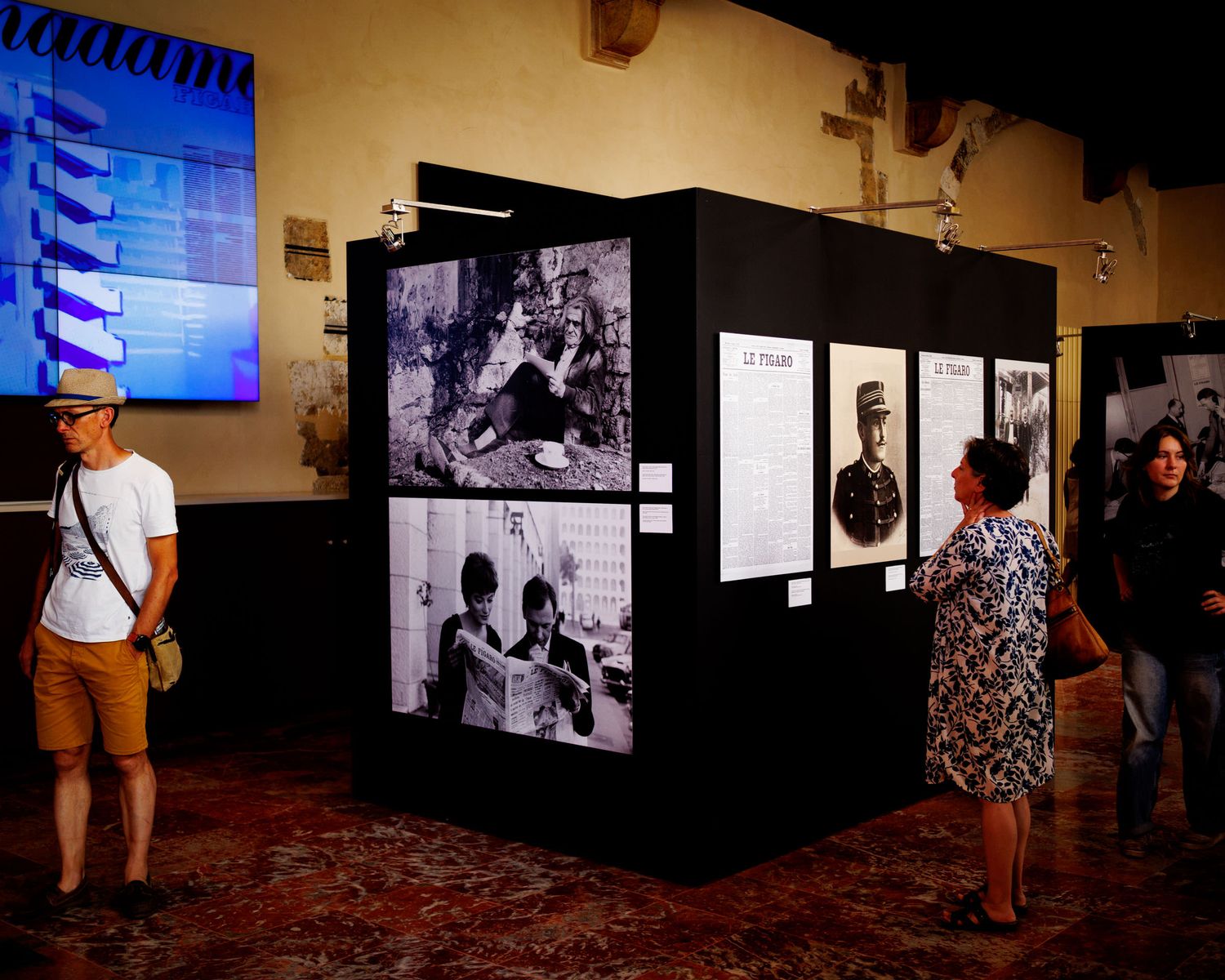Visitors examining black and white photography exhibition displayed on a freestanding cube in a historic hall.