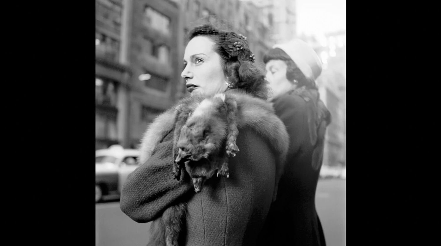 Black and white street photo of a woman in a fur-collared coat gazing sideways on a busy city sidewalk with another person behind.