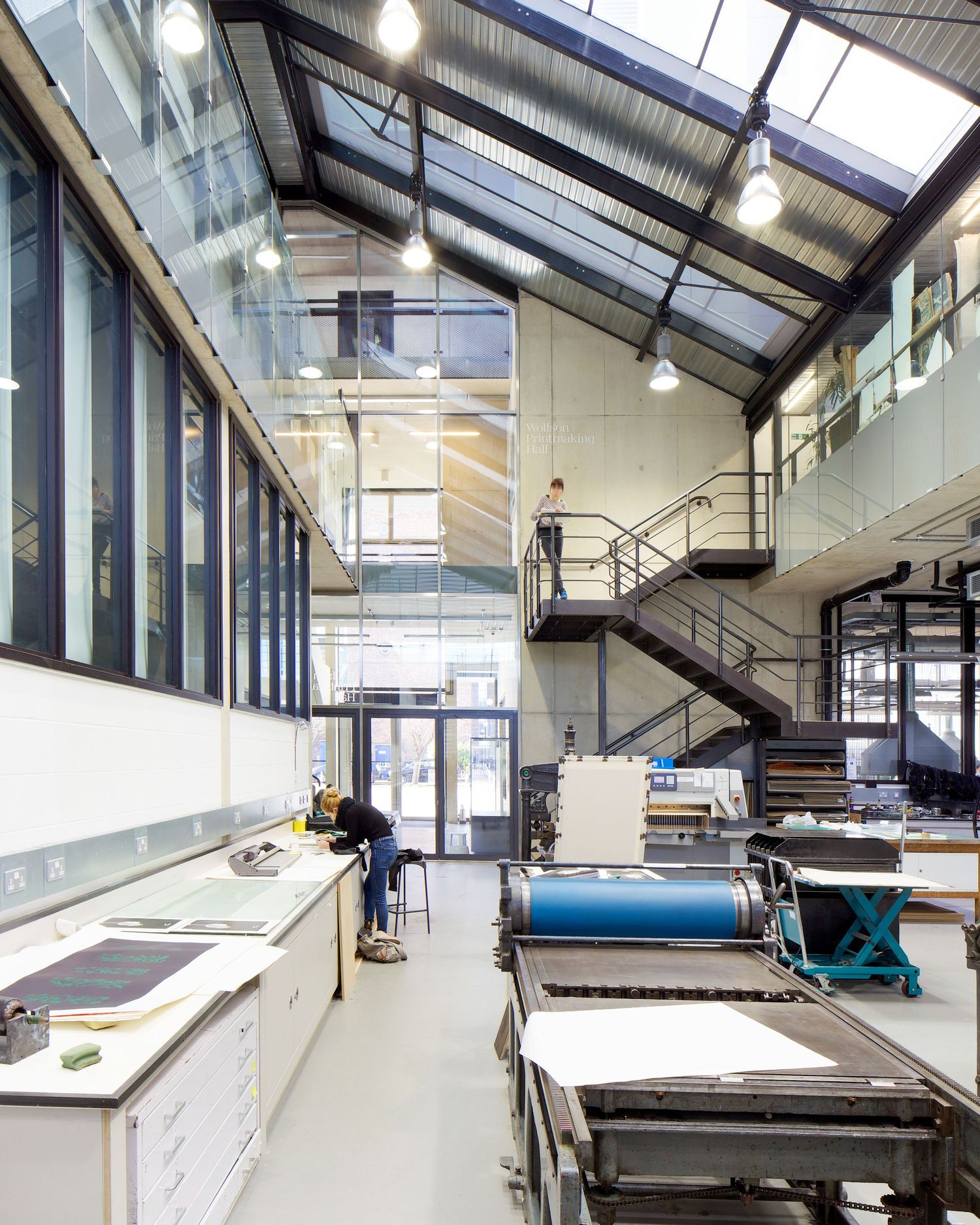 Industrial print workshop with large machinery, skylights, mezzanine level, and workers at workbenches in a modern studio space.