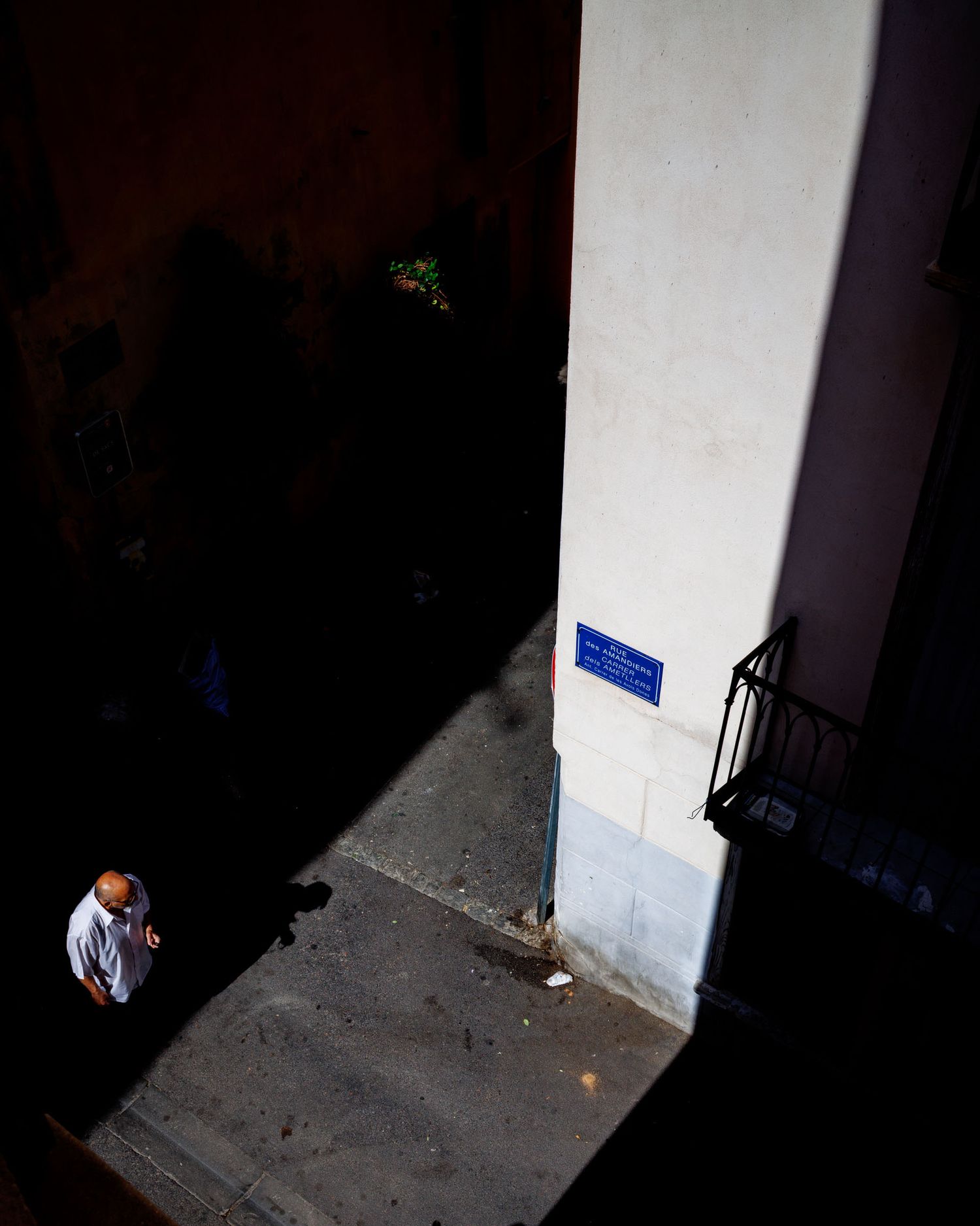 Aerial view of a man walking past a white building corner with a blue street sign in sunlight.